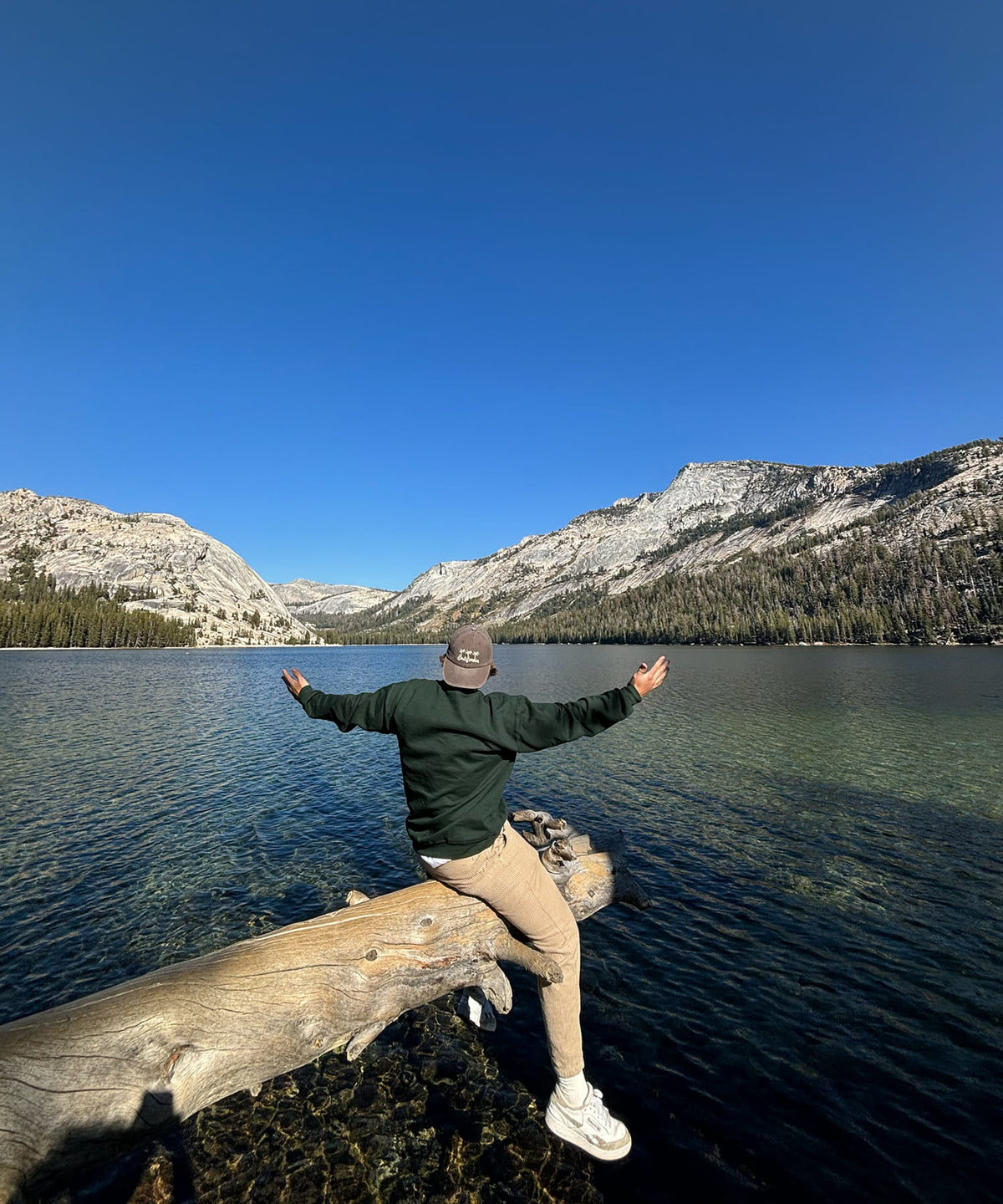 A person sitting on a fallen log by a clear mountain lake, with their arms outstretched, under a bright blue sky. They are wearing a dark green hoodie, beige pants, and white sneakers.