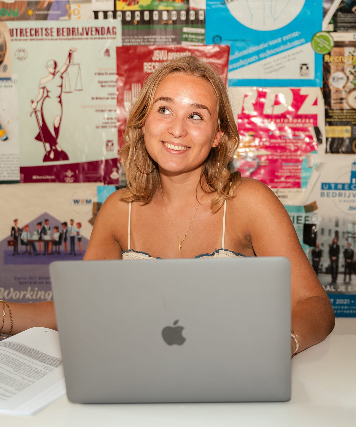 A smiling blonde woman sits at a white desk using a silver Apple MacBook laptop, with a colorful wall of posters and flyers behind her.
